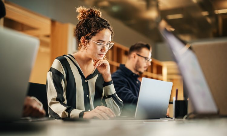 A focused young woman working on a laptop in a shared office space, with other people in the background also engaged in their work, creating a collaborative work environment.