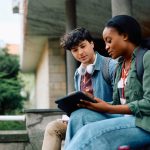 Two college students sitting outside looking at tablet together.