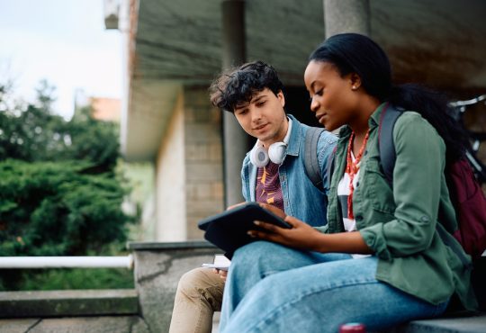 Two college students sitting outside looking at tablet together.