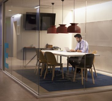 Man sitting alone in an empty glass office working on a laptop.