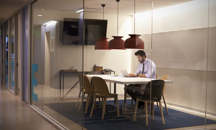 Man sitting alone in an empty glass office working on a laptop.