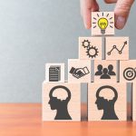 Person stacking wooden blocks on table, with icons of people, handshake, gears, bullseye and lightbulb