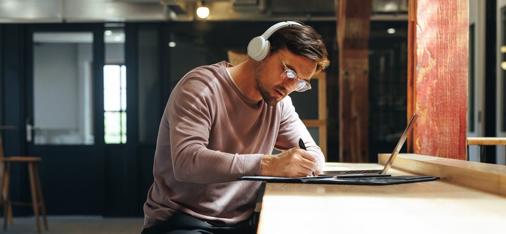 Man sitting at high table writing and wearing over-ear headphones