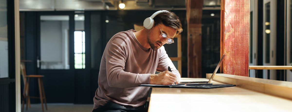 Man sitting at high table writing and wearing over-ear headphones