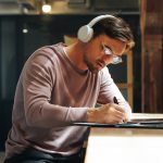 Man sitting at high table writing and wearing over-ear headphones