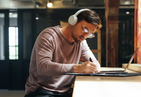 Man sitting at high table writing and wearing over-ear headphones