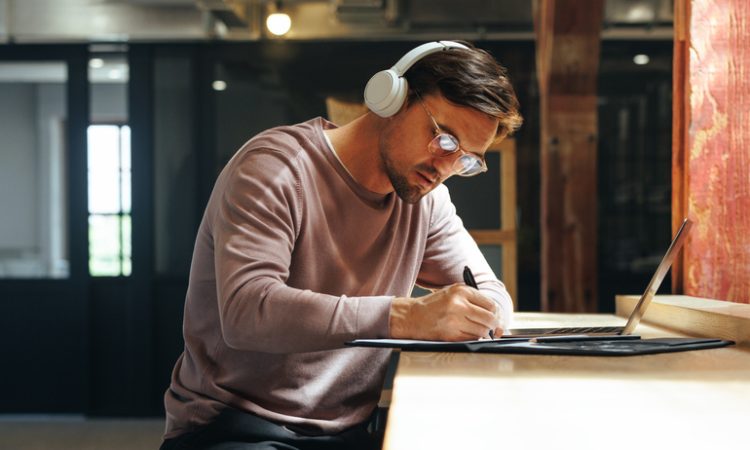 Man sitting at high table writing and wearing over-ear headphones