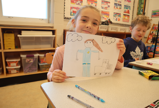 Young girl holding up drawing in classroom.