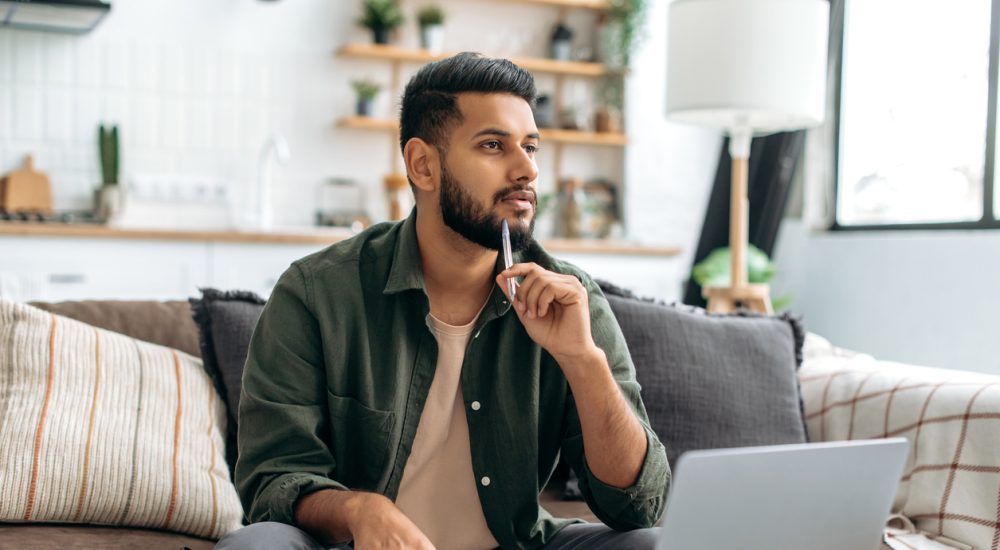 Man thinking with pen resting on chin while sitting in front of laptop