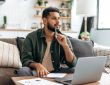 Man thinking with pen resting on chin while sitting in front of laptop