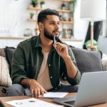 Man thinking with pen resting on chin while sitting in front of laptop