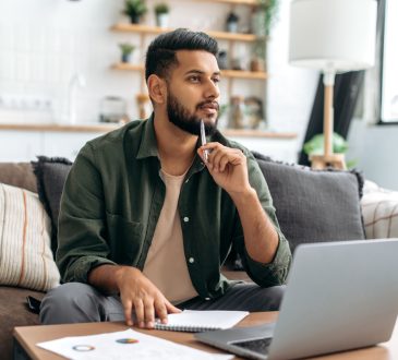 Man thinking with pen resting on chin while sitting in front of laptop