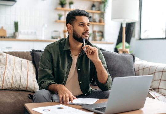 Man thinking with pen resting on chin while sitting in front of laptop