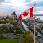 A flowing Canadian flag on a flagpole with Old Quebec City and the St. Lawrence River in the background.