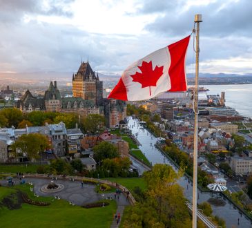 A flowing Canadian flag on a flagpole with Old Quebec City and the St. Lawrence River in the background.