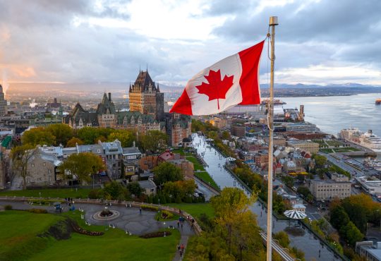 A flowing Canadian flag on a flagpole with Old Quebec City and the St. Lawrence River in the background.