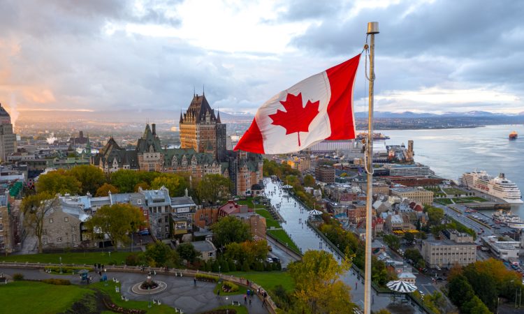 A flowing Canadian flag on a flagpole with Old Quebec City and the St. Lawrence River in the background.