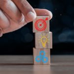 Closeup of hand stacking wooden blocks with icons of bullseye, lightbulb and gears