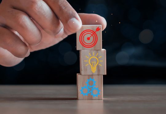 Closeup of hand stacking wooden blocks with icons of bullseye, lightbulb and gears