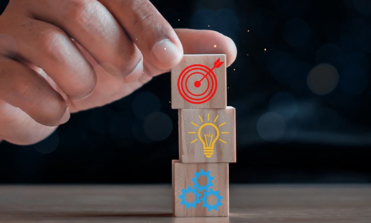 Closeup of hand stacking wooden blocks with icons of bullseye, lightbulb and gears