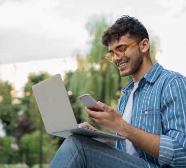 Young man sitting outside with computer balanced on lap, looking at phone in hand