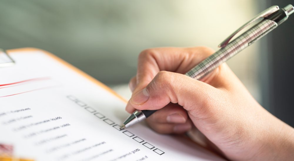 Hand ticking off checkmarks on list with pen attached to clipboard