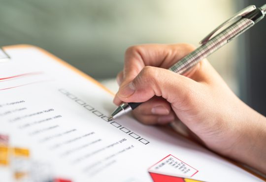 Hand ticking off checkmarks on list with pen attached to clipboard