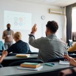 Rear view of man raising arm to ask a question during a presentation in lecture hall.