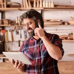 Smiling man with a rugged beard, talking on his mobile phone while looking at a digital tablet in his hand, standing in his woodwork studio