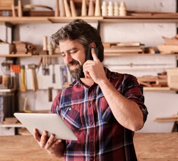 Smiling man with a rugged beard, talking on his mobile phone while looking at a digital tablet in his hand, standing in his woodwork studio