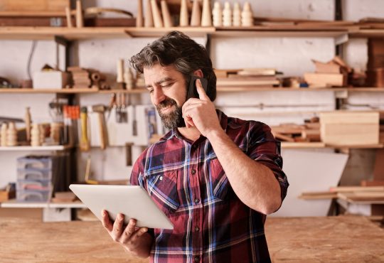 Smiling man with a rugged beard, talking on his mobile phone while looking at a digital tablet in his hand, standing in his woodwork studio