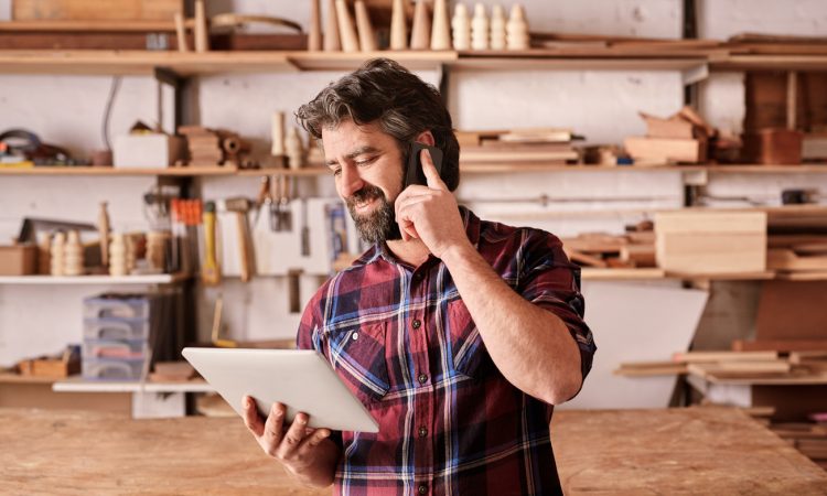 Smiling man with a rugged beard, talking on his mobile phone while looking at a digital tablet in his hand, standing in his woodwork studio