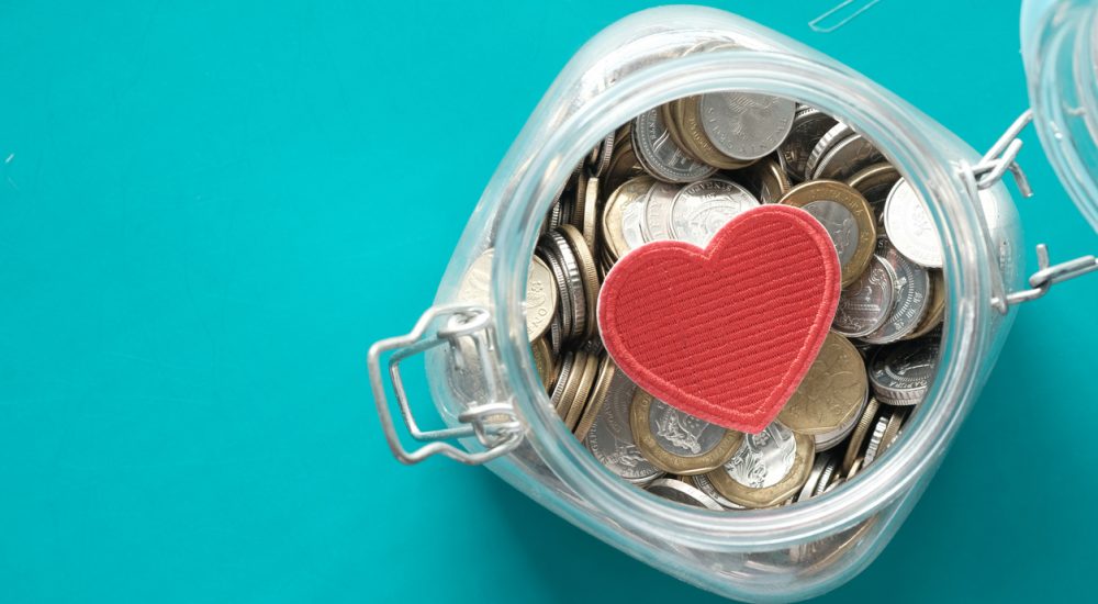 Red heart and coins in a jar on blue background .