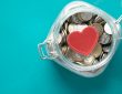 Red heart and coins in a jar on blue background .