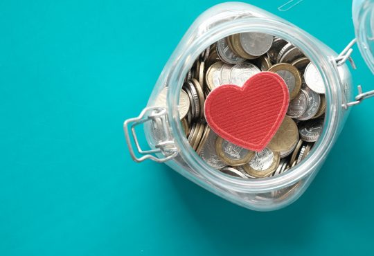 Red heart and coins in a jar on blue background .