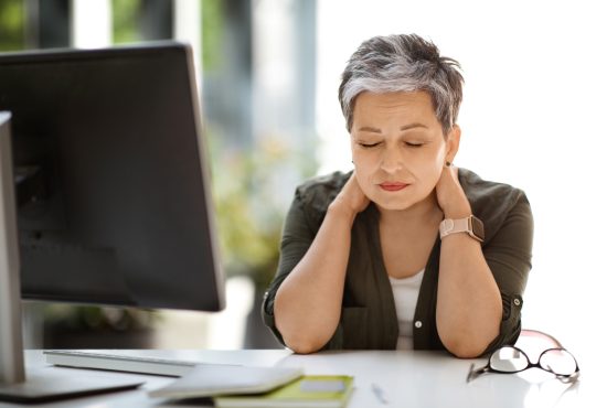 Woman sitting at desk with eyes closed, rubbing neck