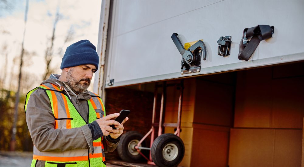 Delivery truck driver standing behind box, checking phone