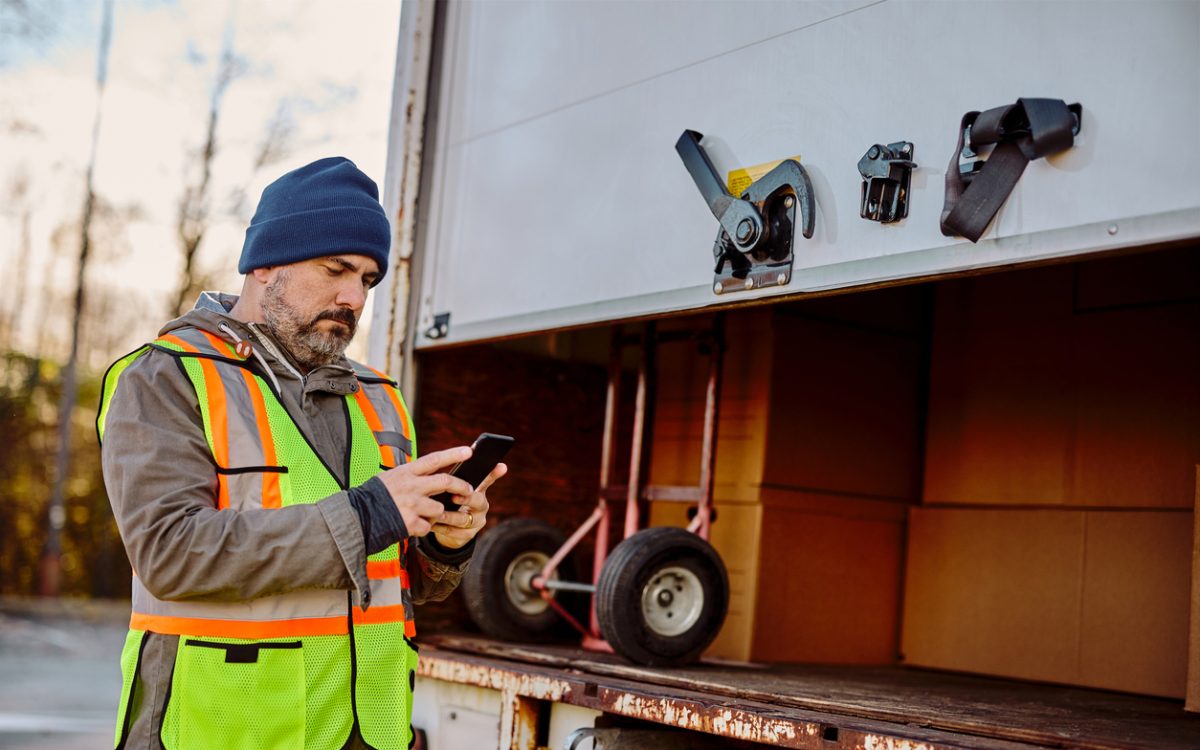 Delivery truck driver standing behind box, checking phone