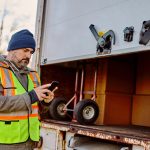 Delivery truck driver standing behind box, checking phone
