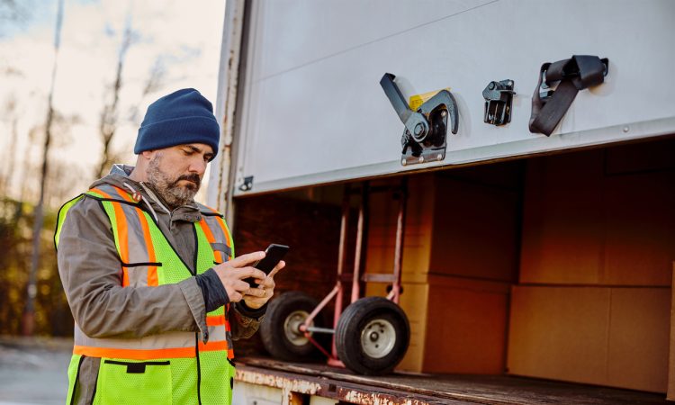 Delivery truck driver standing behind box, checking phone