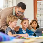 Male teacher helping schoolchildren in a research work using digital tablet in classroom. Three elementary school pupils working on digital tablet at desk while talking to teacher. Classmates in class using technology for studying with the assistance of teacher at elementary school.