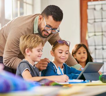 Male teacher helping schoolchildren in a research work using digital tablet in classroom. Three elementary school pupils working on digital tablet at desk while talking to teacher. Classmates in class using technology for studying with the assistance of teacher at elementary school.