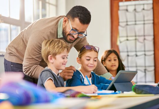 Male teacher helping schoolchildren in a research work using digital tablet in classroom. Three elementary school pupils working on digital tablet at desk while talking to teacher. Classmates in class using technology for studying with the assistance of teacher at elementary school.