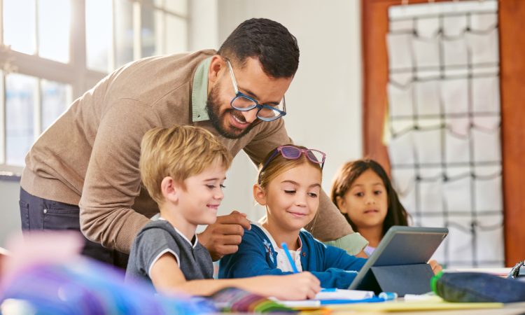 Male teacher helping schoolchildren in a research work using digital tablet in classroom. Three elementary school pupils working on digital tablet at desk while talking to teacher. Classmates in class using technology for studying with the assistance of teacher at elementary school.