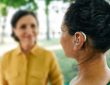 Adult woman with a hearing impairment uses a hearing aid to communicate with her female friend at city park.