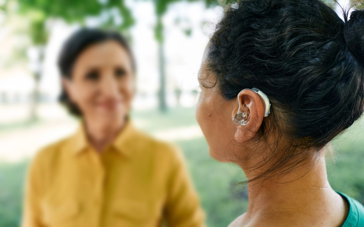Adult woman with a hearing impairment uses a hearing aid to communicate with her female friend at city park.