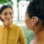 Adult woman with a hearing impairment uses a hearing aid to communicate with her female friend at city park.