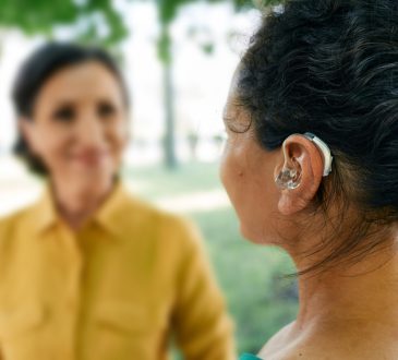 Adult woman with a hearing impairment uses a hearing aid to communicate with her female friend at city park.