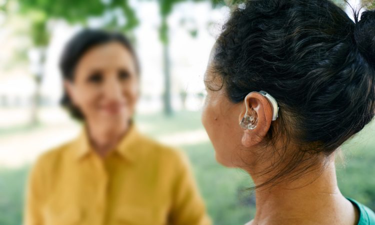 Adult woman with a hearing impairment uses a hearing aid to communicate with her female friend at city park.