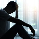 Man silhouetted sitting beside window with head in hands